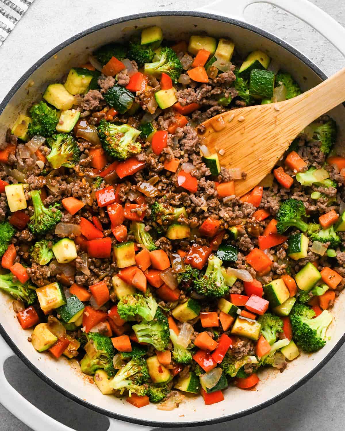 making ground beef stir fry - ground beef and veggies cooked in a white skillet with a black rim and a wooden spatula