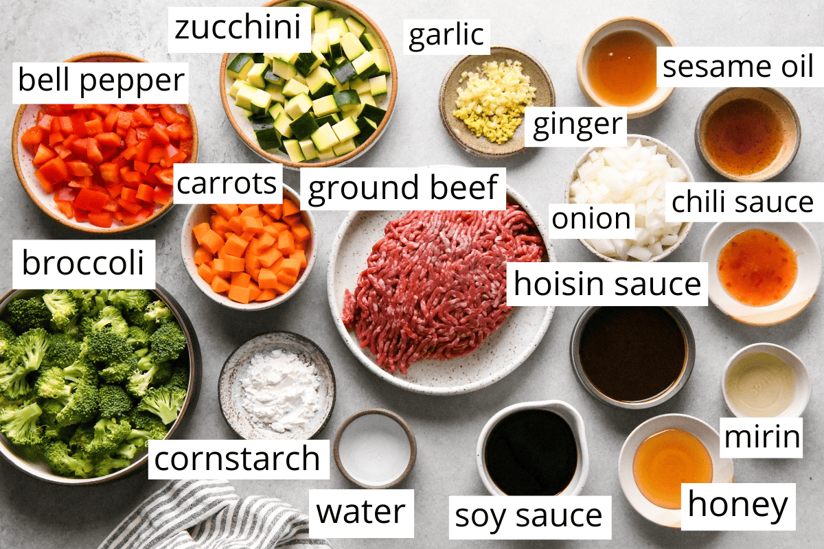 overhead photo of the ingredients in this ground beef stir fry recipe like zucchini, bell pepper, ginger, carrots, sesame oil, ground beef, soy sauce and more all in bowls and labeled