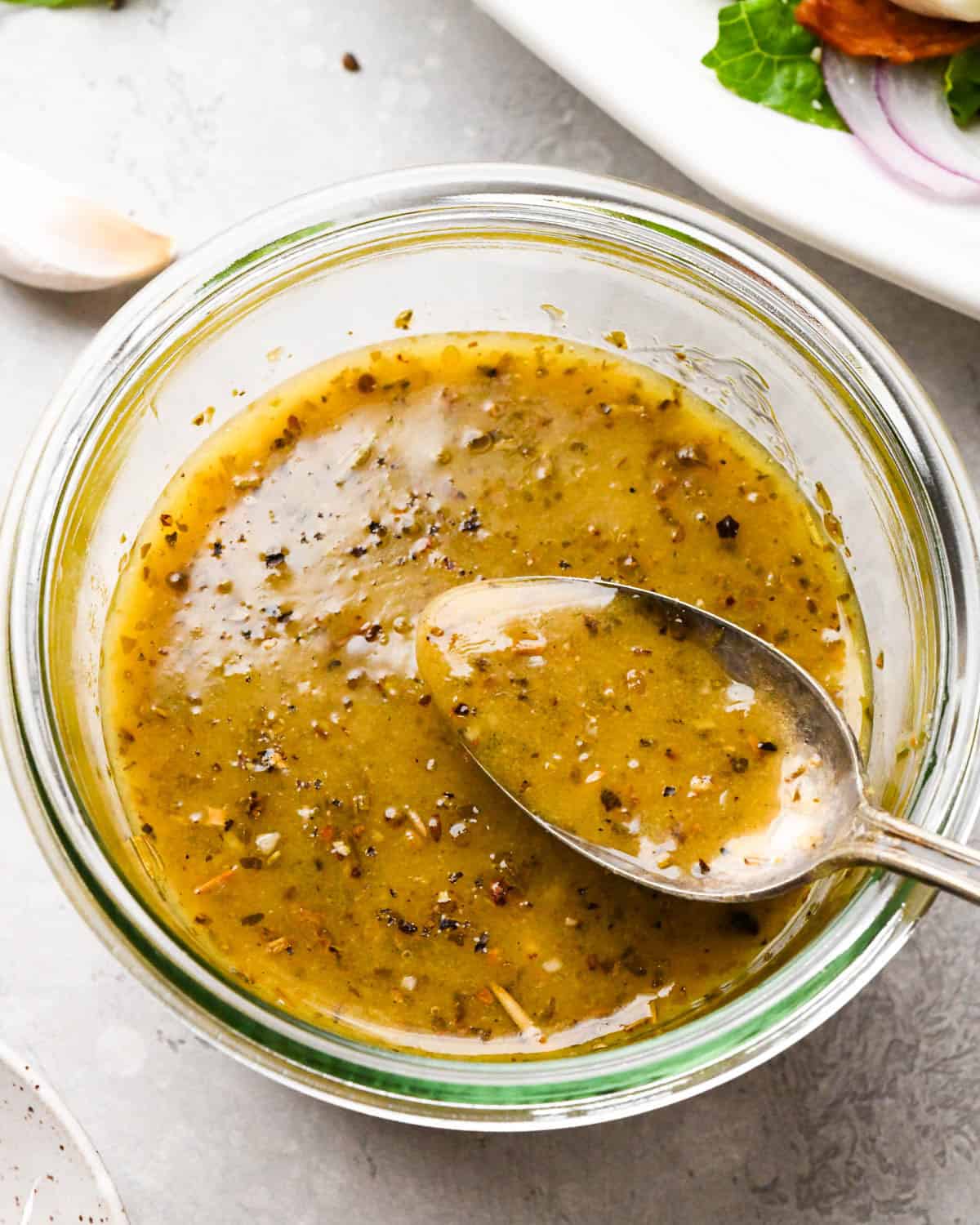 Italian dressing recipe in a glass jar with a spoon hovering over the jar slightly with dressing on it, with garlic and a salad in the background