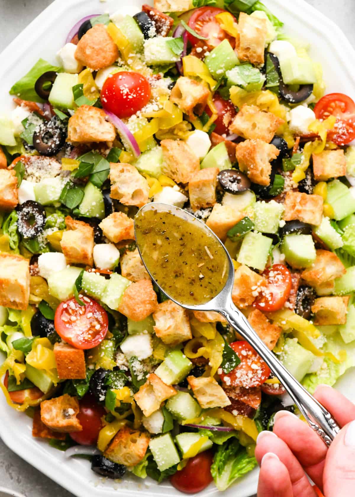 Italian dressing on a silver spoon about to be poured over an italian salad with croutons, cucumbers, black olives, etc.