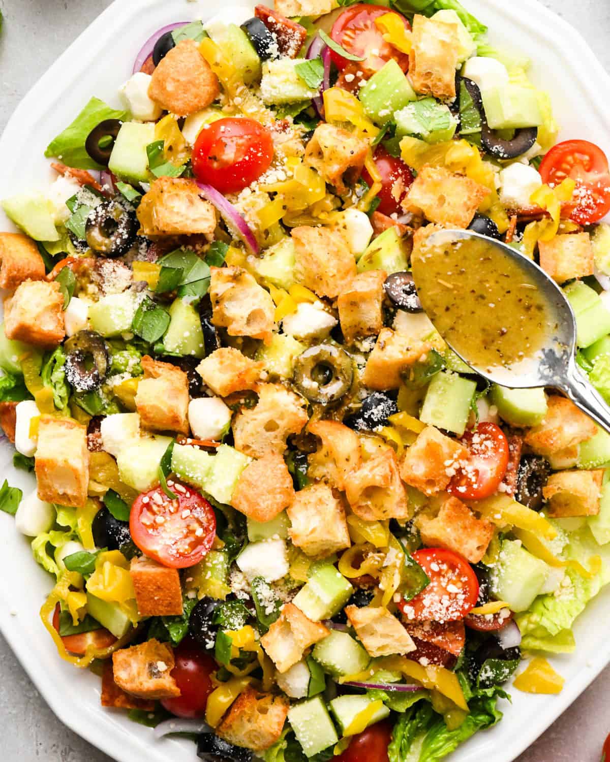Italian dressing on a silver spoon about to be poured over an italian salad with croutons, cucumbers, black olives, etc.