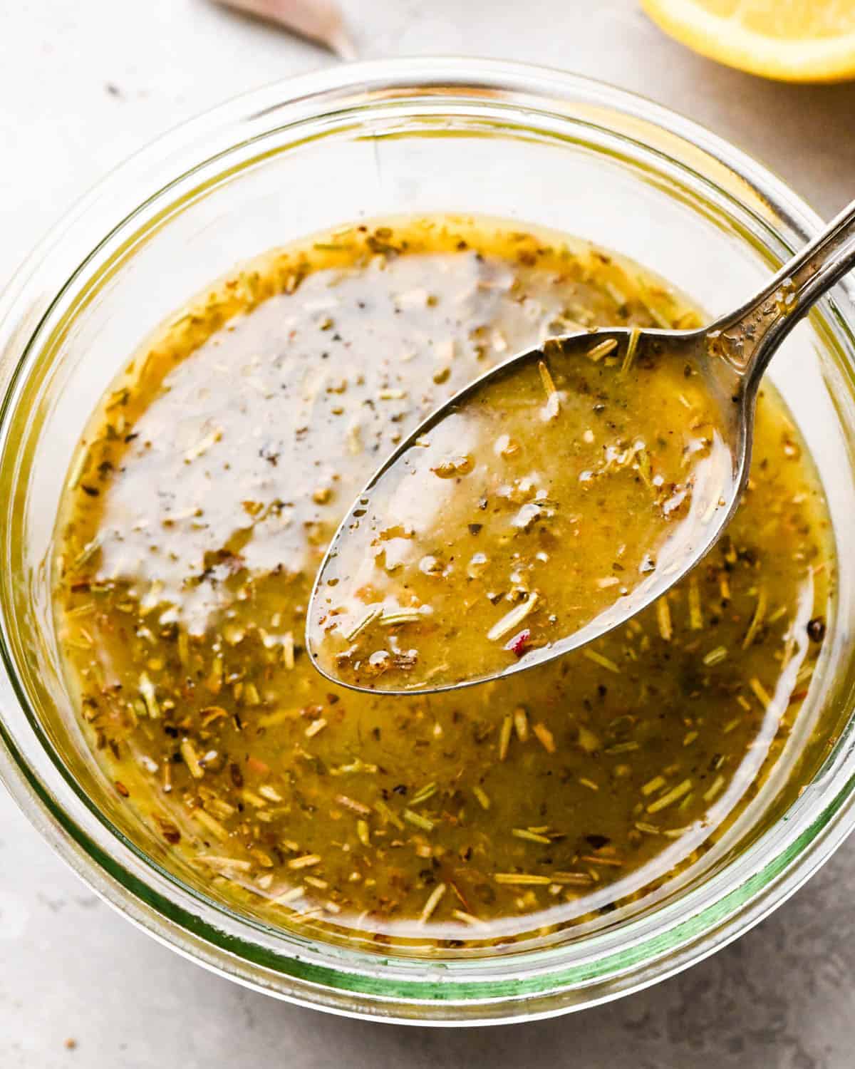 Italian dressing recipe in a glass jar with a spoon hovering over the dressing with dressing on it, with a lemon in the background