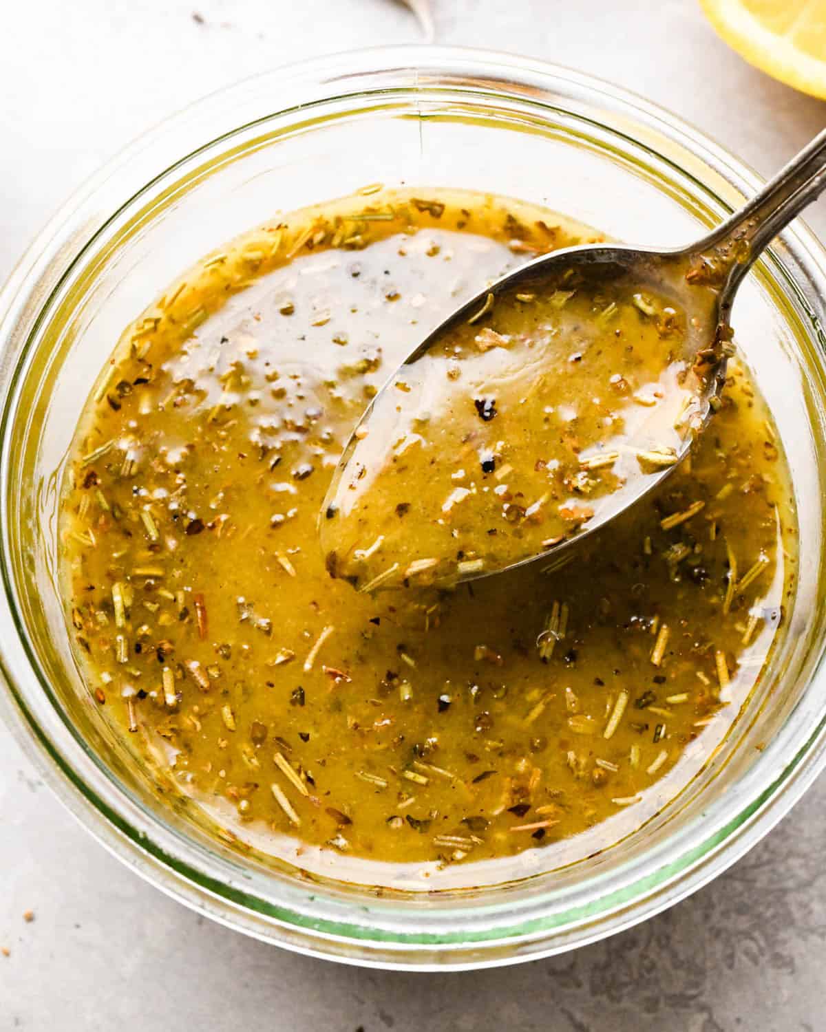 Italian dressing recipe in a glass jar with a spoon hovering over the dressing with dressing on it, with a lemon in the background