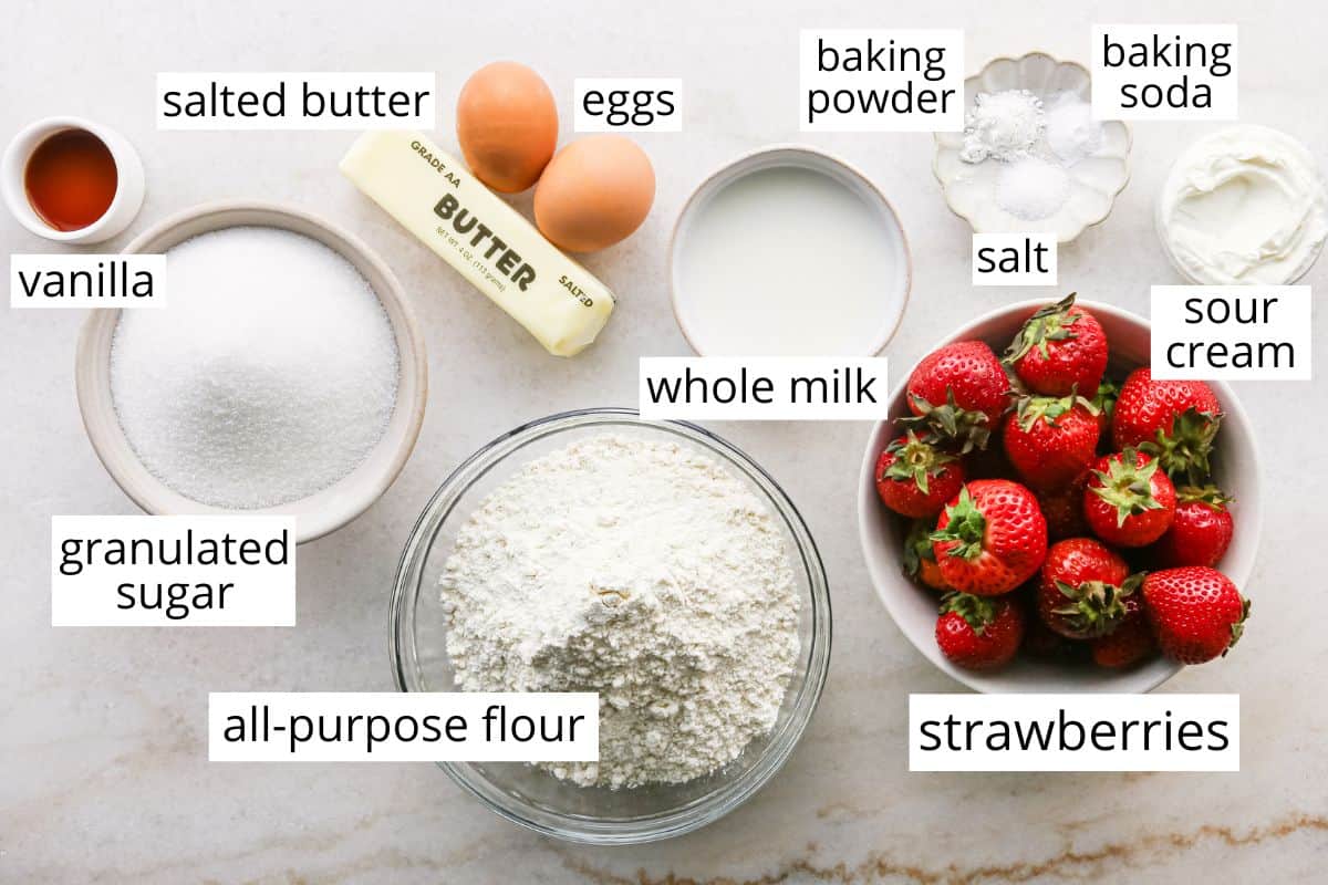 overhead photo of the ingredients in this strawberry cake recipe in bowls and labeled like sugar, flour, milk, butter, eggs, and strawberries.