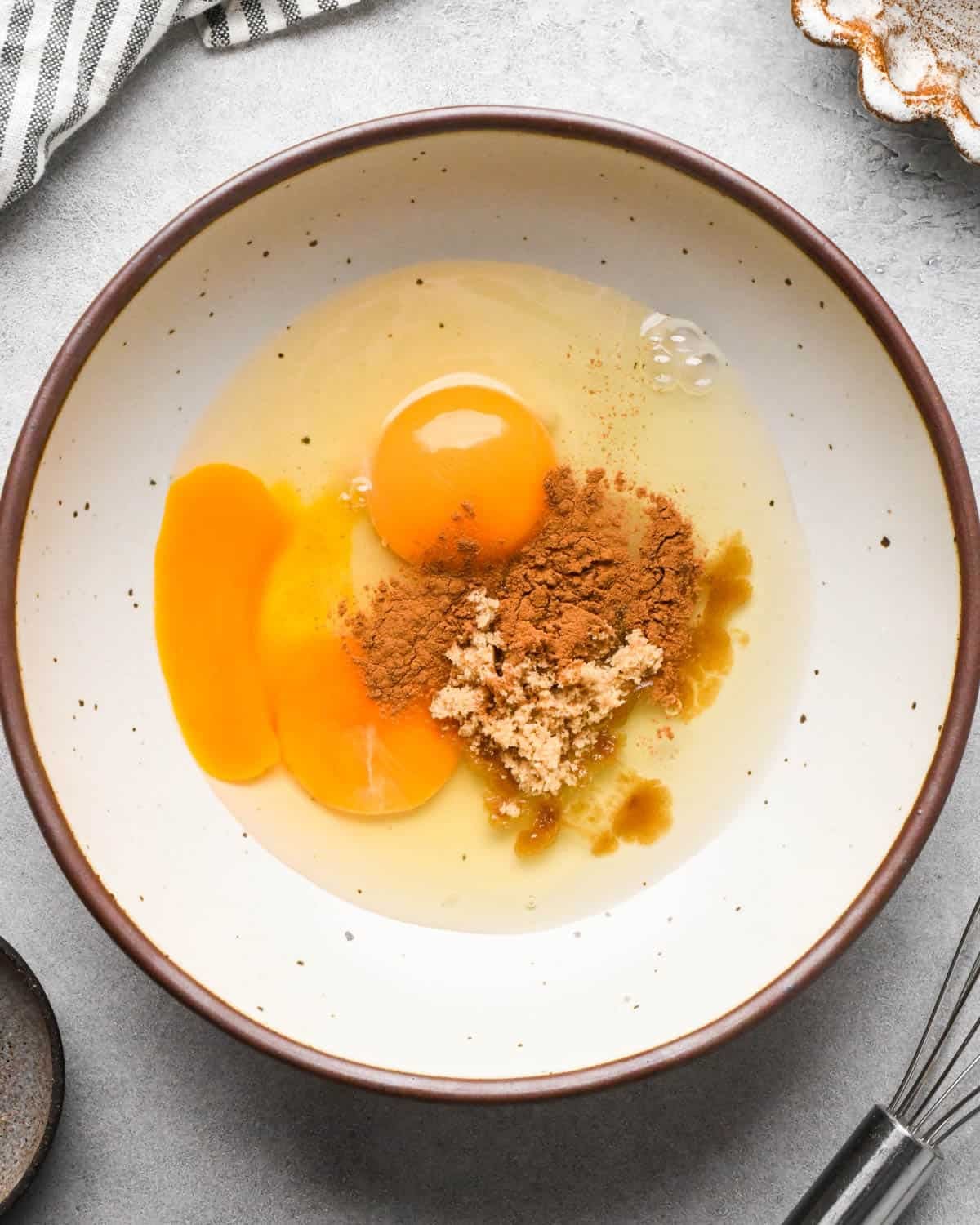 Making banana bread French toast: overhead photo of eggs, cinnamon, brown sugar, and vanilla sitting together in a white speckled bowl on a gray countertop.