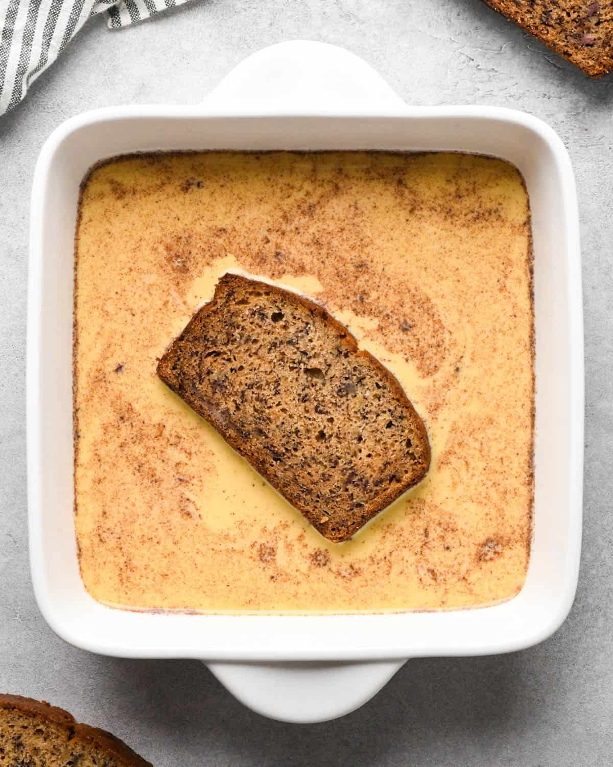Making banana bread French toast: overhead photo of one slice of banana bread being soaked in an egg mixture in a white square casserole dish.
