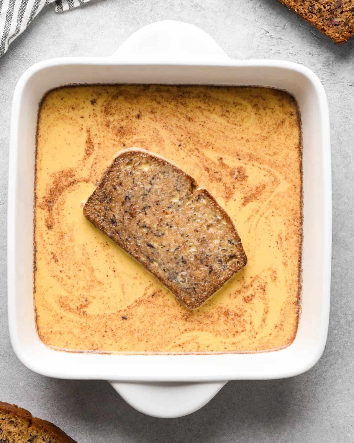 Making banana bread French toast: overhead photo of one slice of banana bread being soaked in an egg mixture in a white square casserole dish.