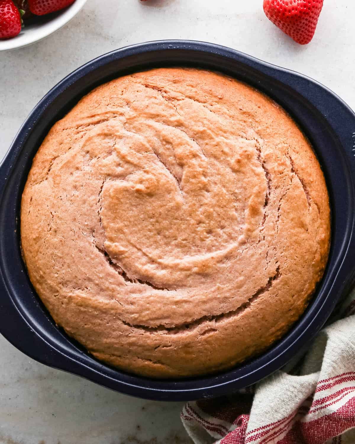 baked strawberry cake in a round, navy blue cake pan with strawberries and a red and cream striped towel around it.