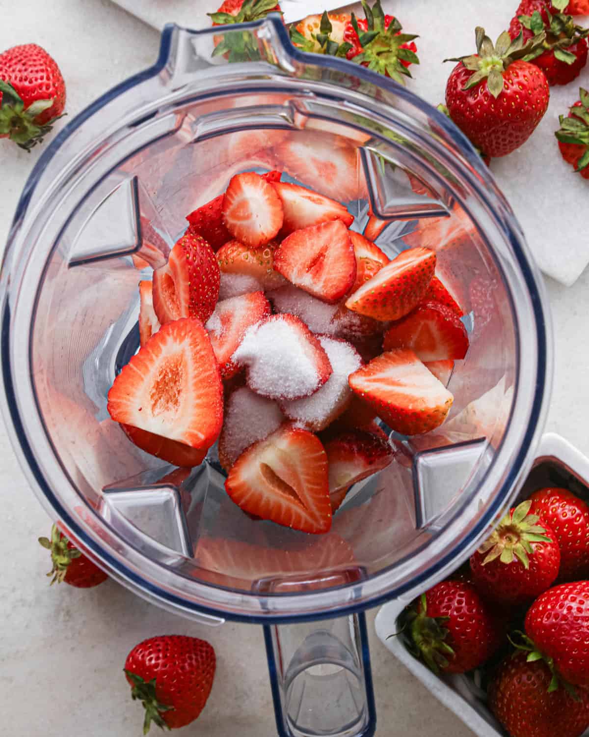 making strawberry cake - halved strawberries and sugar in a blending container, surrounded by strawberries