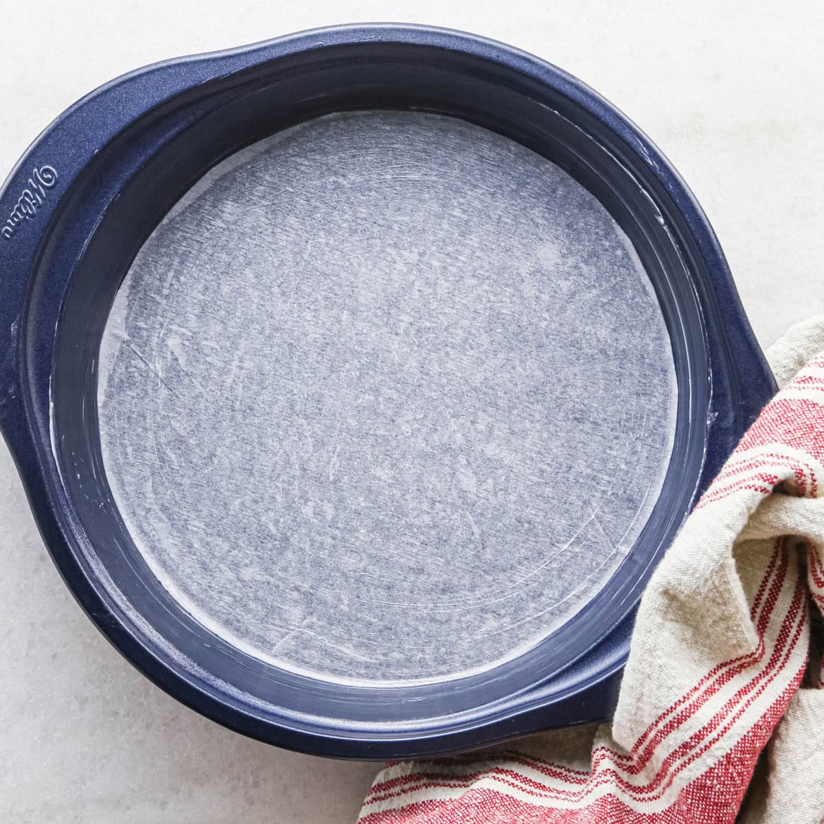 making strawberry cake - a 9" round, blue cake plate lined with a circle of parchment paper. there is a red and cream striped towel in the bottom right corner