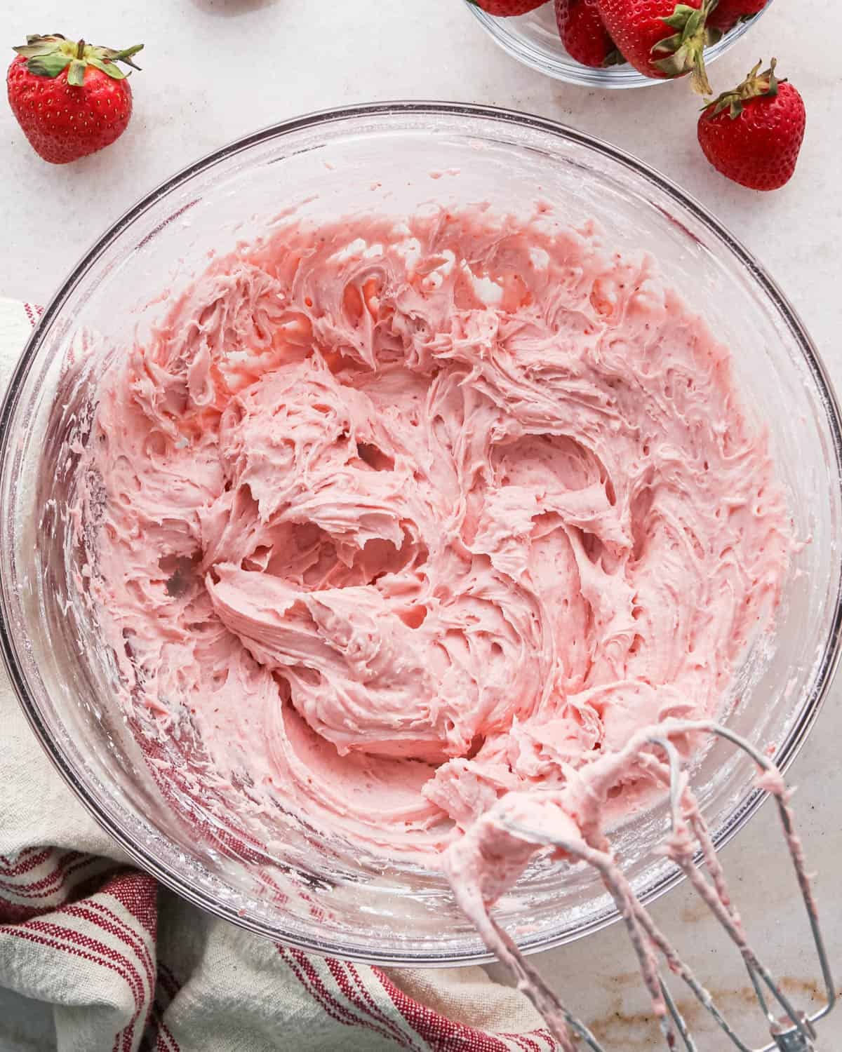 strawberry frosting in a glass bowl with two beaters setting on the side of the bowl, surrounded by strawberries and a red and cream striped towel