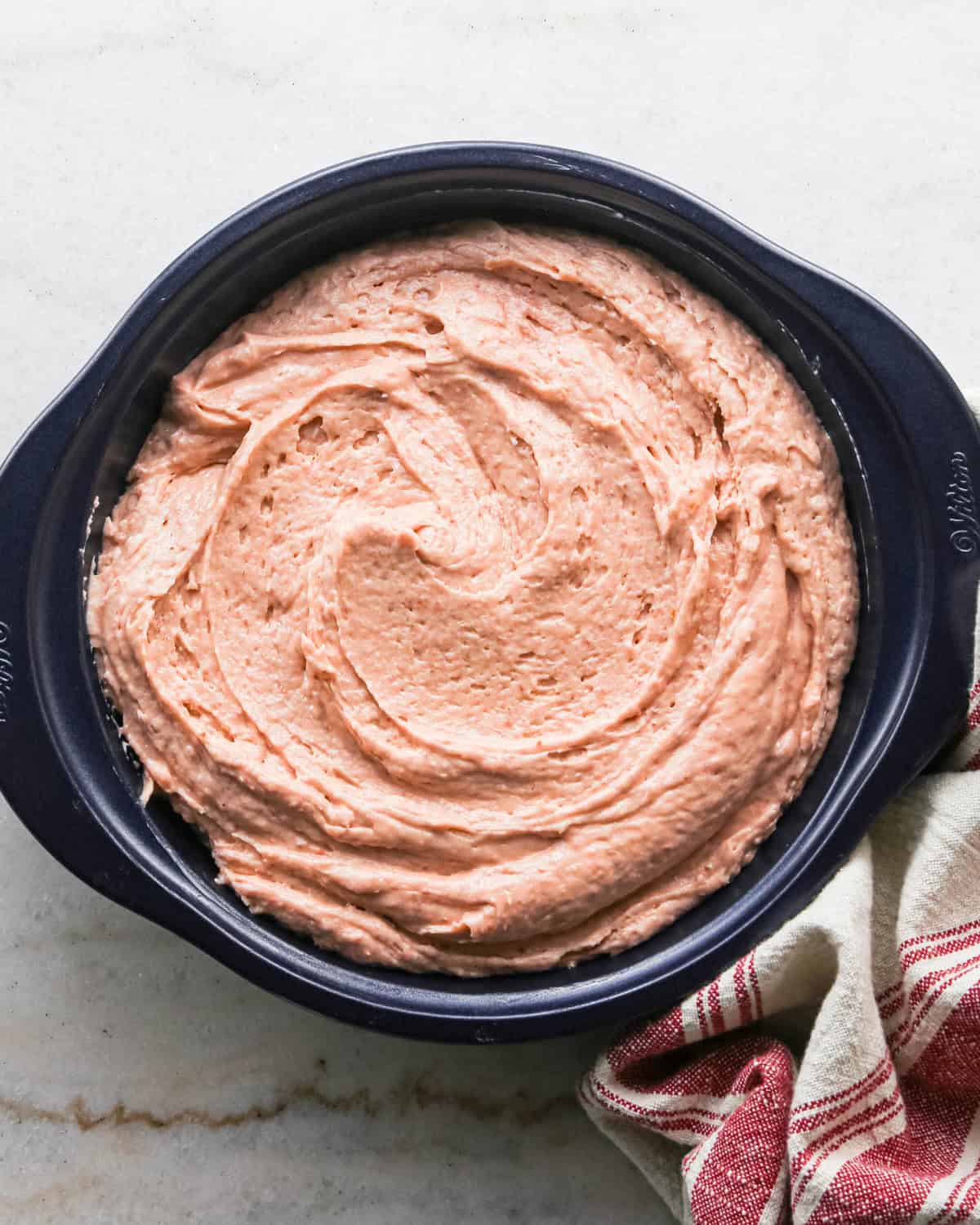 strawberry cake batter spread into a round, navy blue cake pan before baking. there is a red and cream striped towel in the bottom right corner