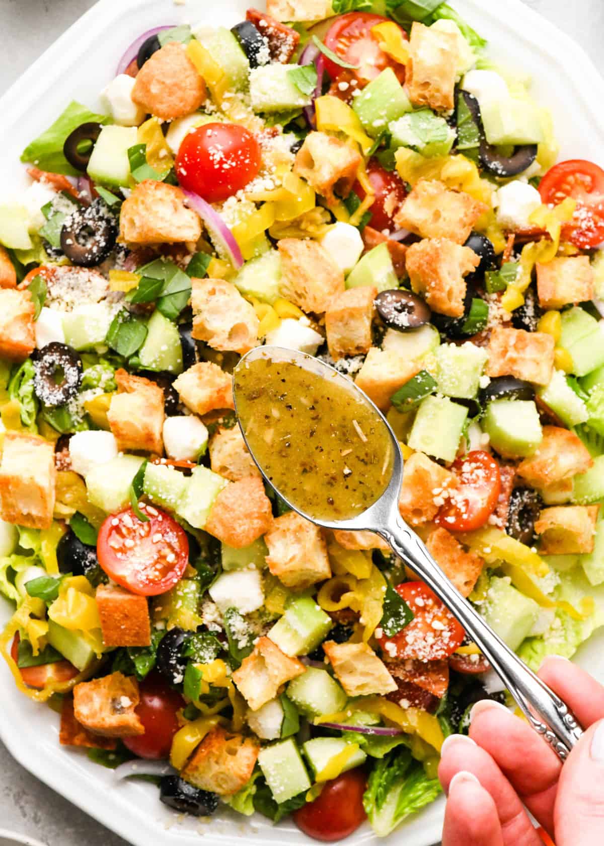 a spoon of Italian dressing being held over an assembled Italian salad ready to drizzle on top