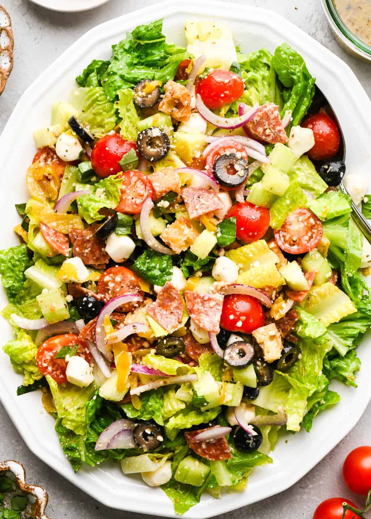 Italian salad on a white serving tray with a silver serving spoon surrounded by tomatoes and dressing