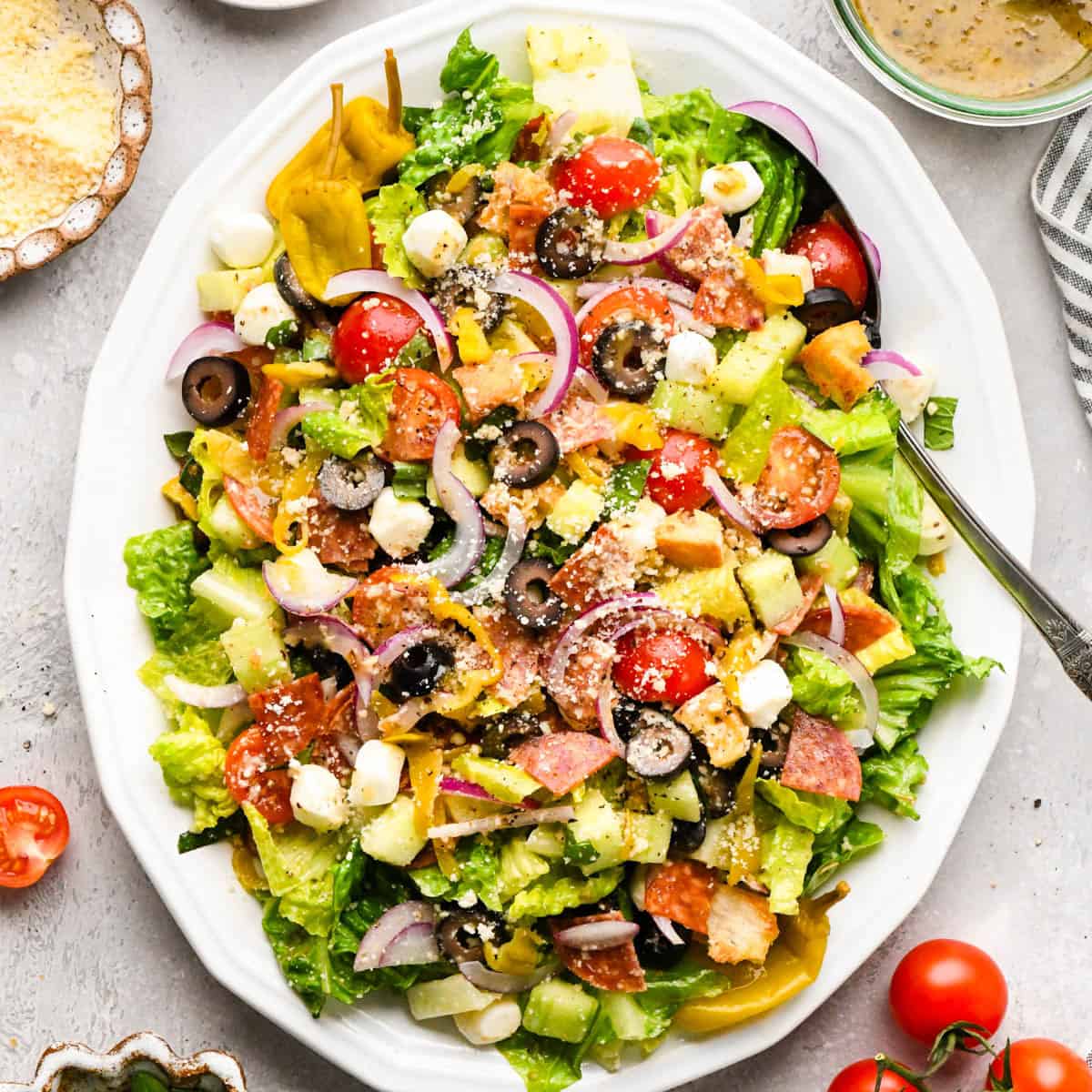 Italian salad on a white serving tray with a silver serving spoon surrounded by parmesan cheese, tomatoes and dressing