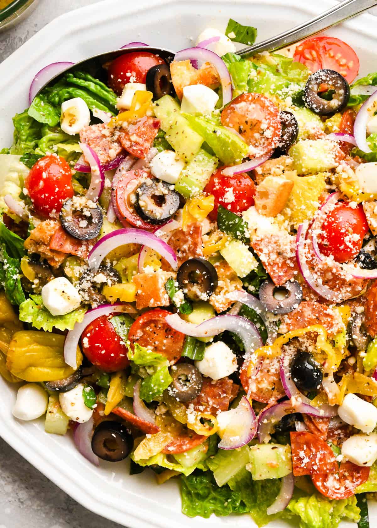 close up photo of Italian salad with black olives, cucumbers, tomatoes, lettuce, basil, and croutons on a serving plate with a silver spoon