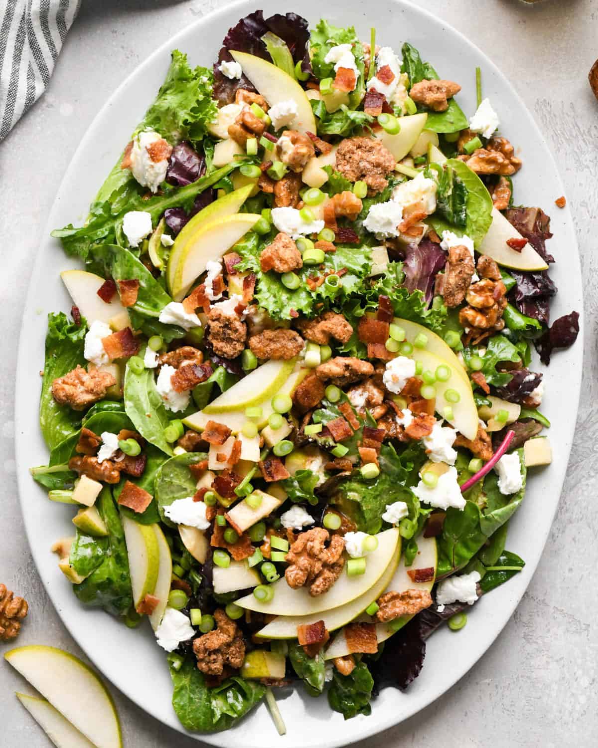 Pear salad with sliced pears, bacon, goat cheese, candied walnuts, green onions, and mixed greens, tossed in a homemade balsamic vinaigrette on a white serving platter.