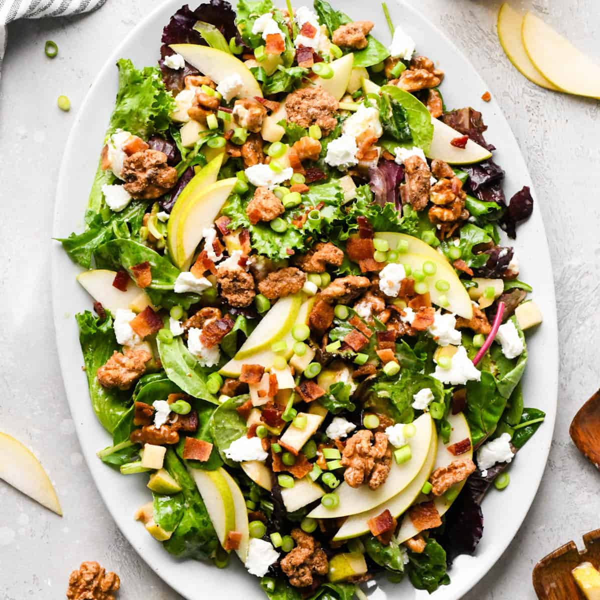 Pear salad with sliced pears, bacon, goat cheese, candied walnuts, green onions, and mixed greens, tossed in a homemade balsamic vinaigrette on a white serving platter. on a gray surface with pears and other items in the background