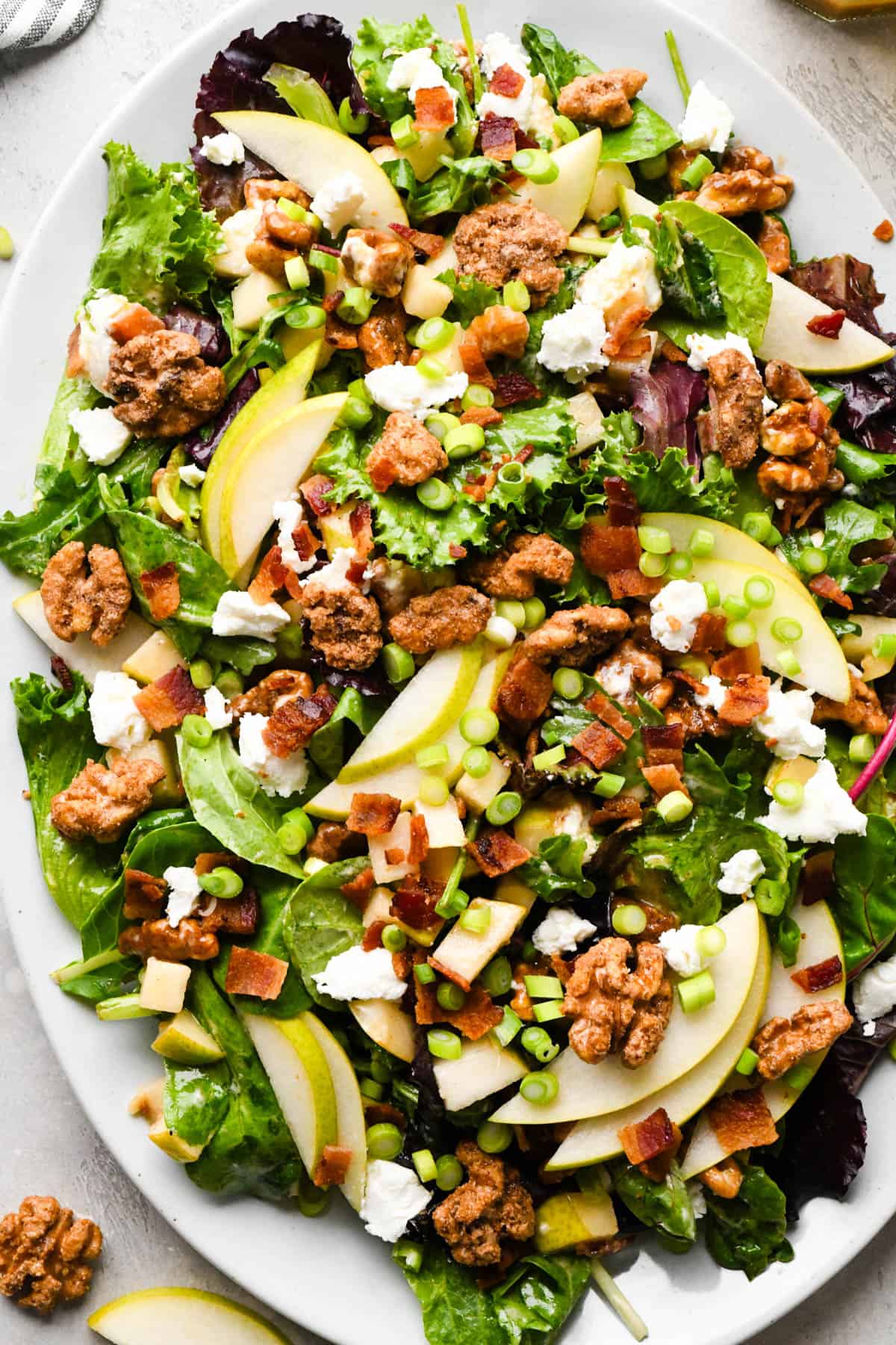 Pear salad with sliced pears, bacon, goat cheese, candied walnuts, green onions, and mixed greens, tossed in a homemade balsamic vinaigrette on a white serving platter.