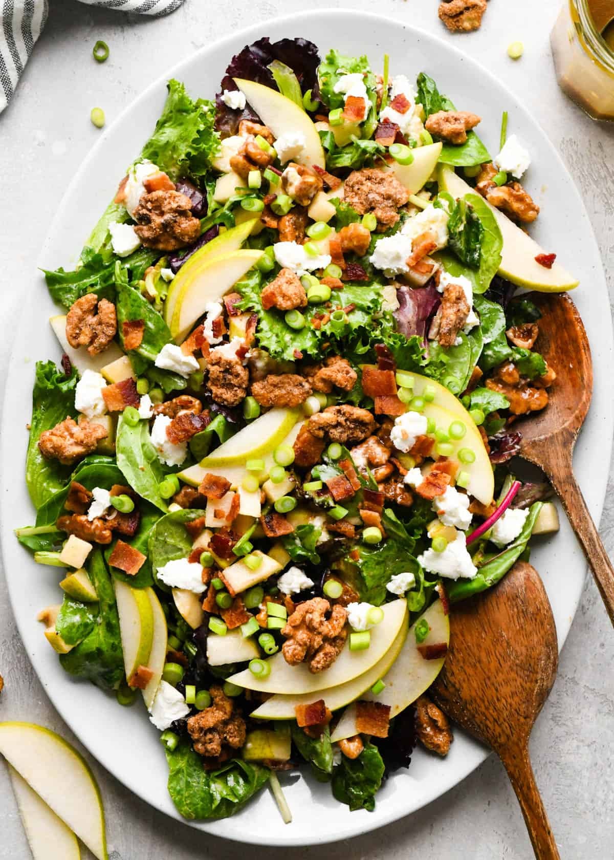 Pear salad with sliced pears, bacon, goat cheese, candied walnuts, green onions, and mixed greens, tossed in a homemade balsamic vinaigrette on a white serving platter with two wooden salad servers