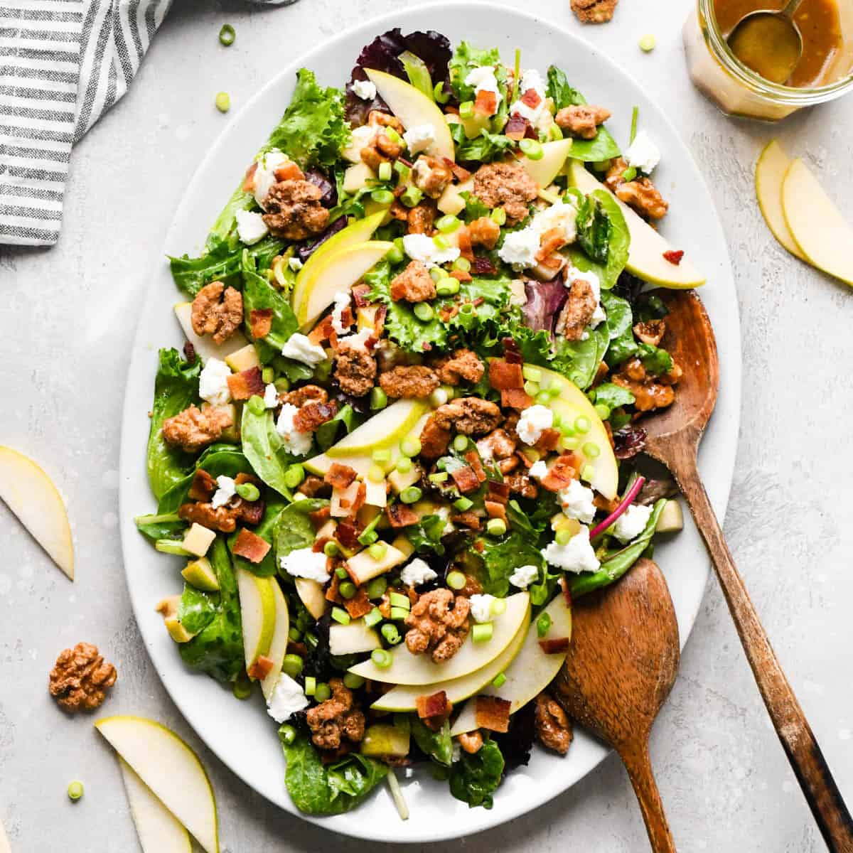 Pear salad with sliced pears, bacon, goat cheese, candied walnuts, green onions, and mixed greens, tossed in a homemade balsamic vinaigrette on a white serving platter with two wooden salad servers on a gray surface