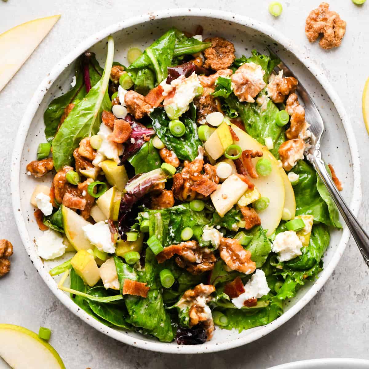 a single serving of Pear salad with sliced pears, bacon, goat cheese, candied walnuts, green onions, and mixed greens, tossed in a homemade balsamic vinaigrette in a white pottery bowl with a silver fork on a gray surface