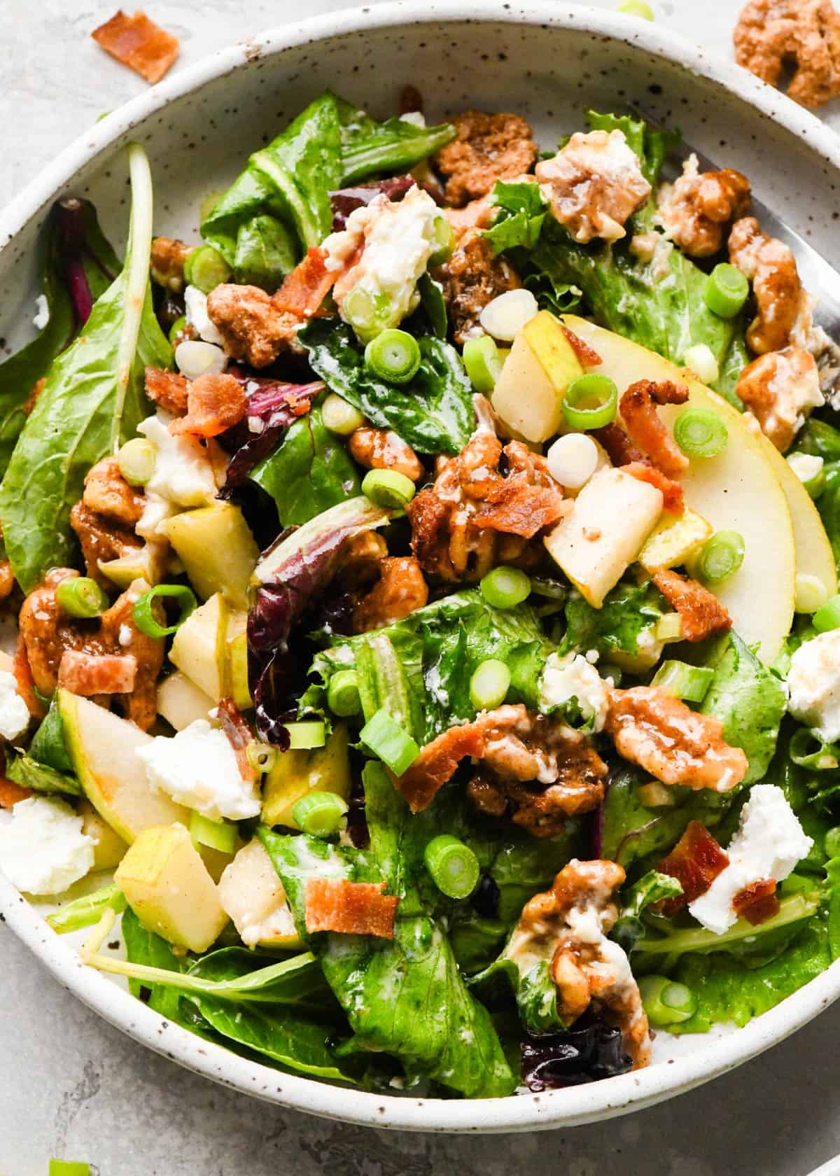 a single serving of Pear salad with sliced pears, bacon, goat cheese, candied walnuts, green onions, and mixed greens, tossed in a homemade balsamic vinaigrette in a white pottery bowl
