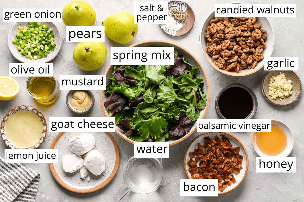 overhead photo of the ingredients for this pear salad recipe labeled and in bowls like spring mix, pears, candied walnuts, goat cheese, bacon and dressing ingredients