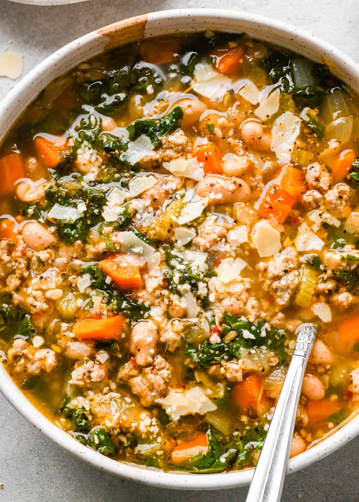 close up photo of sausage, kale and white bean soup in a white ceramic bowl topped with parmesan cheese with a silver spoon, on a gray surface