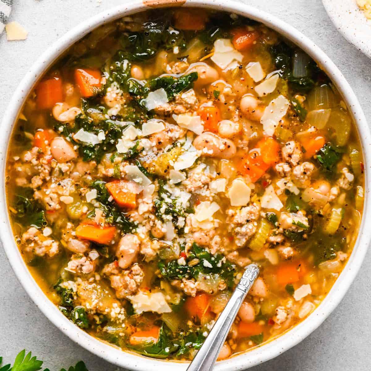 sausage, kale and white bean soup in a white ceramic bowl topped with parmesan cheese with a silver spoon on a gray surface