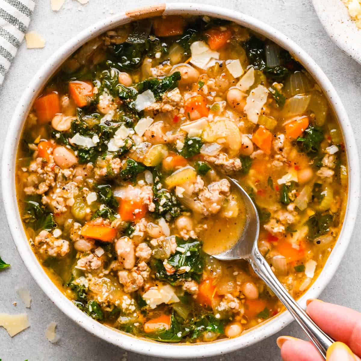 a spoon taking a scoop of sausage, kale and white bean soup in a white ceramic bowl topped with parmesan cheese on a gray surface