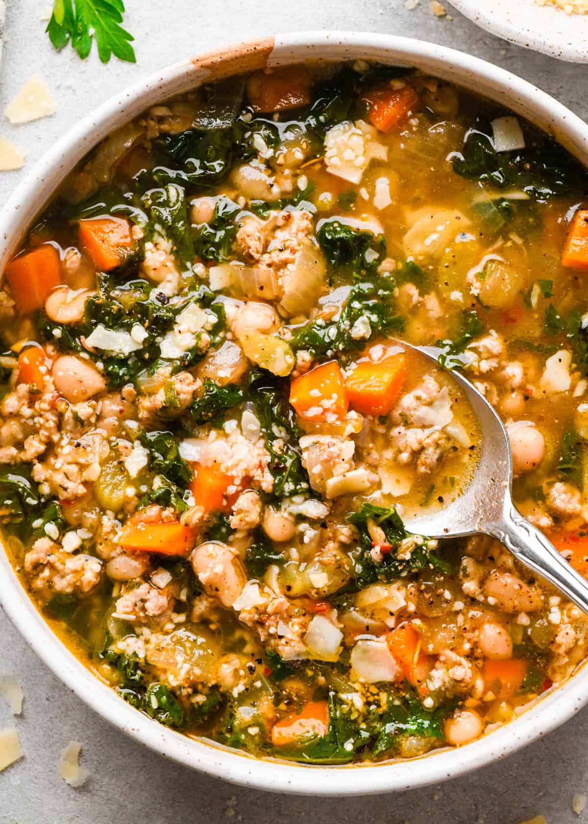 close up photo of a spoon taking a scoop of sausage, kale and white bean soup in a white ceramic bowl topped with parmesan cheese on a gray surface