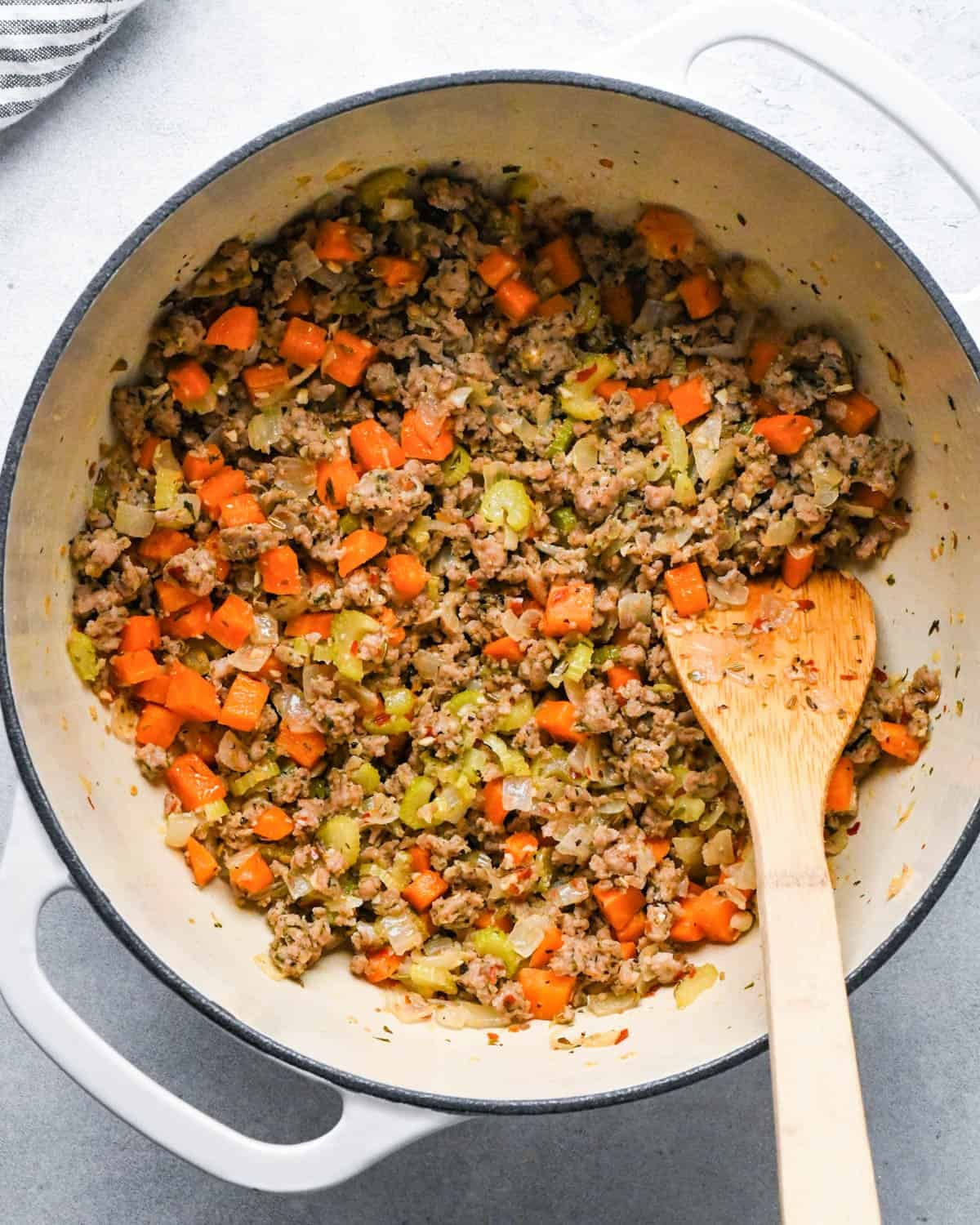 making sausage kale and white bean soup - italian sausage, carrots, celery, onion and garlic in a white pot with a black rim after cooking with a wooden spatula