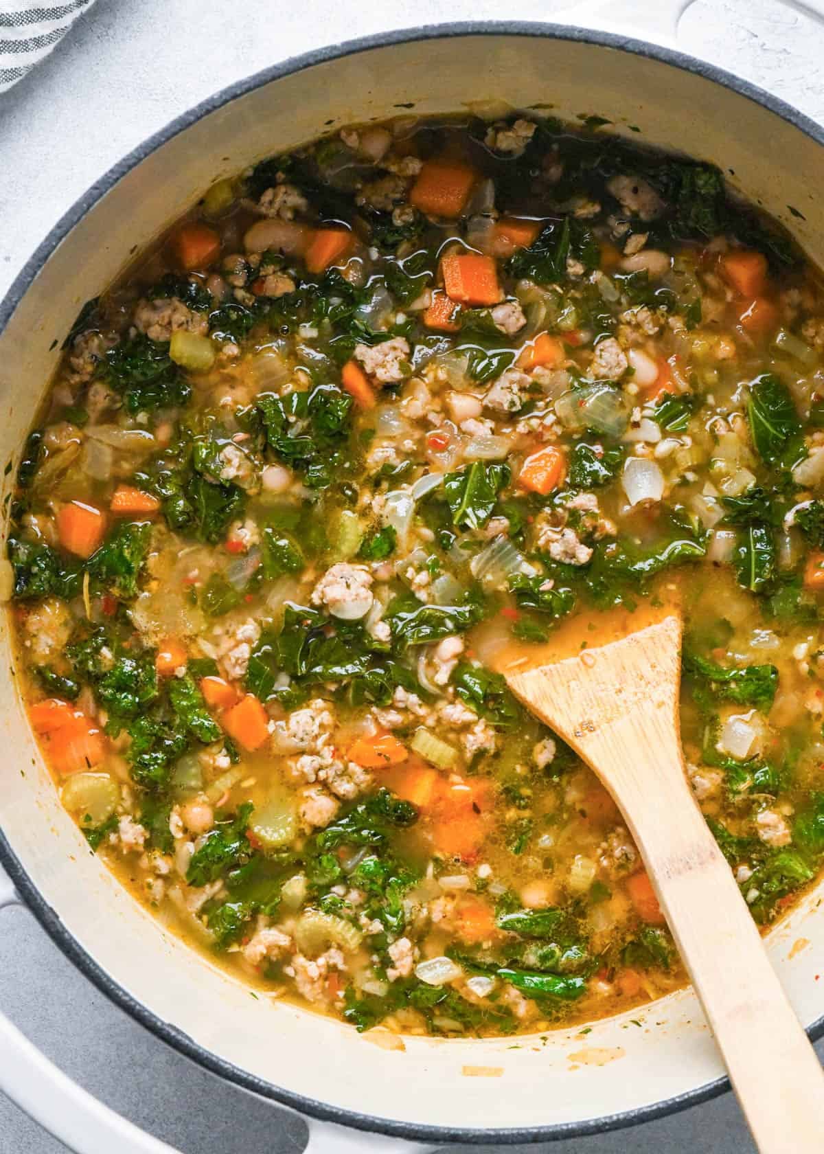 kale and white bean sausage soup in a pot with a wooden spatula