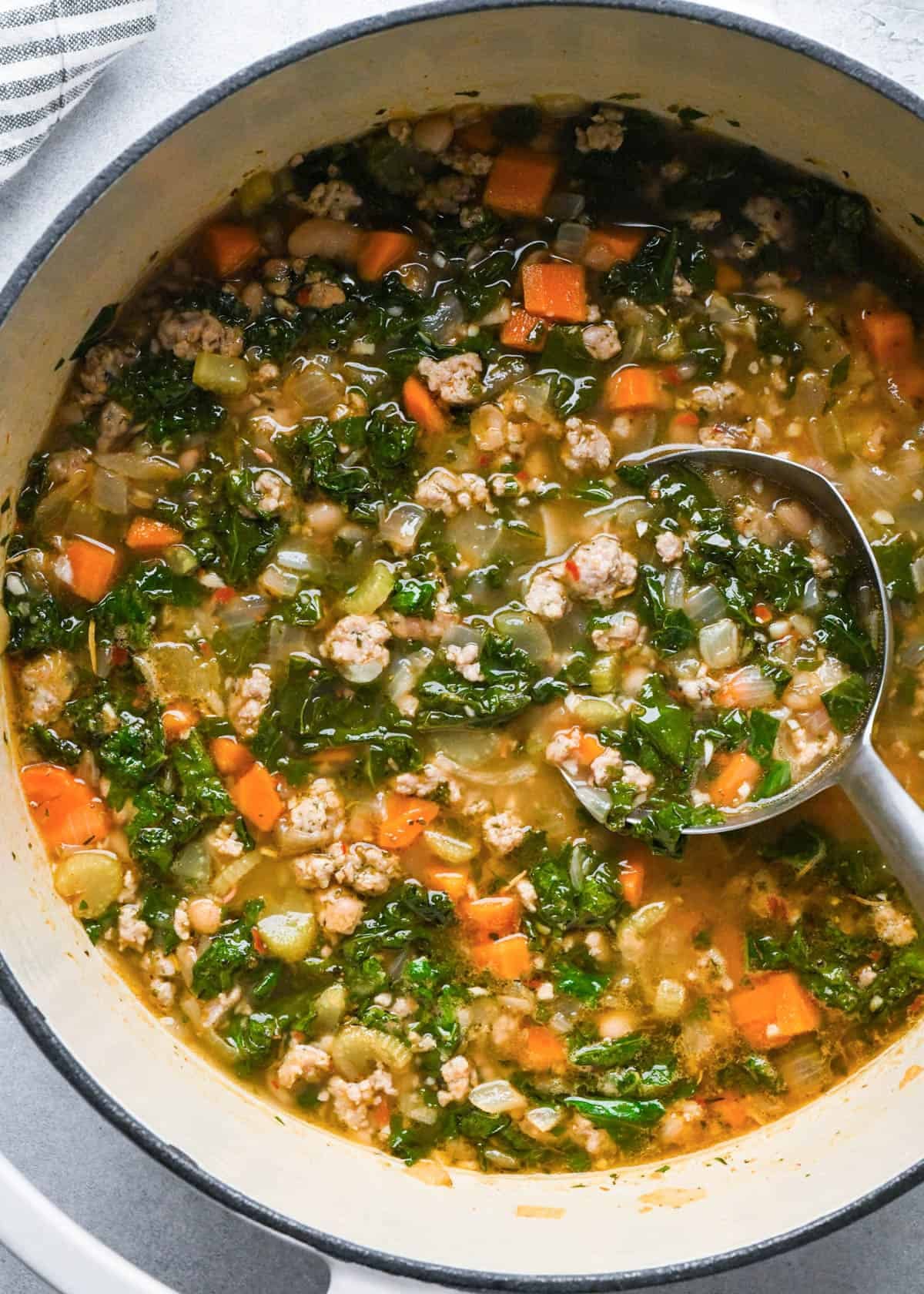 kale and white bean sausage soup in a pot with a ladle spoon taking a scoop of soup