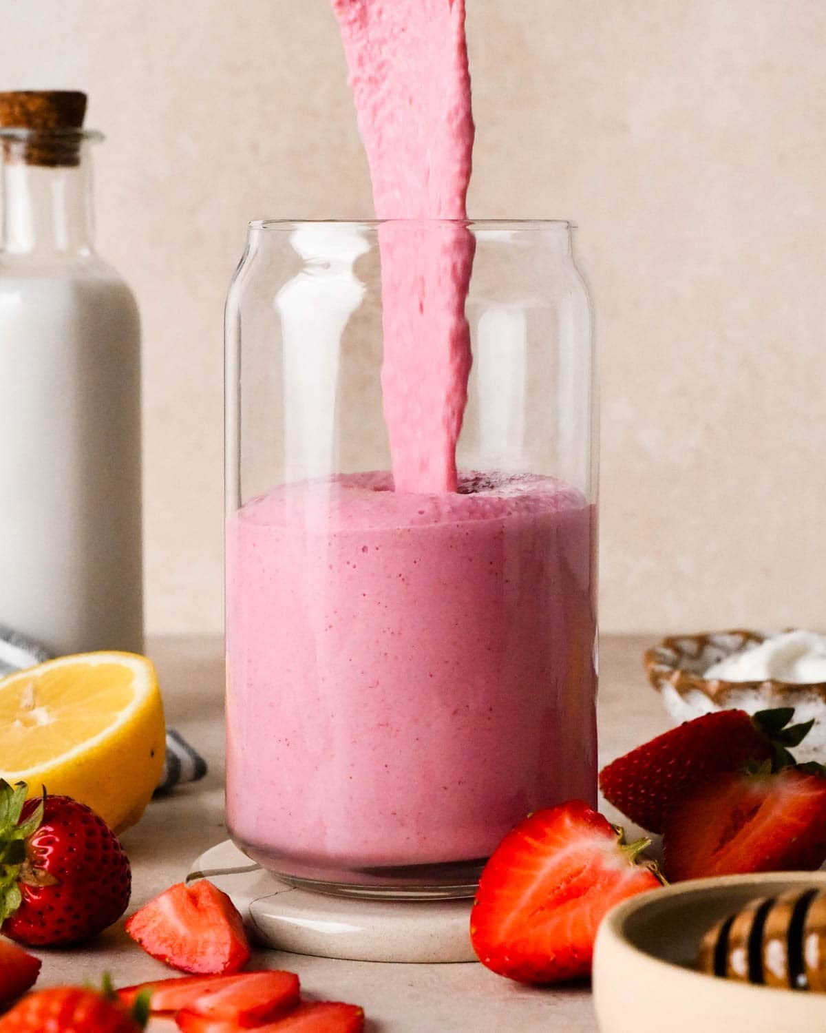 strawberry smoothie being poured from the blending container into a glass, surrounded by strawberries, a half of a lemon, and a bottle of milk in the background