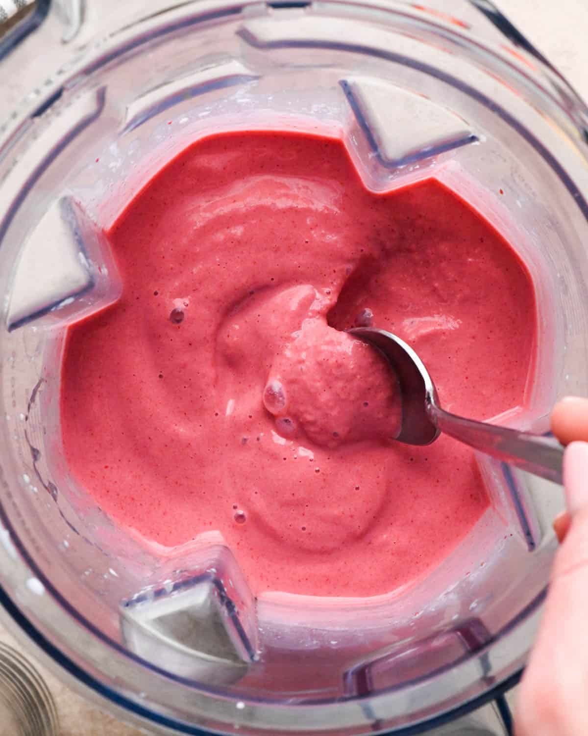 blended strawberry smoothie in a blending container with a spoon scooping some out to show it's thick creamy texture