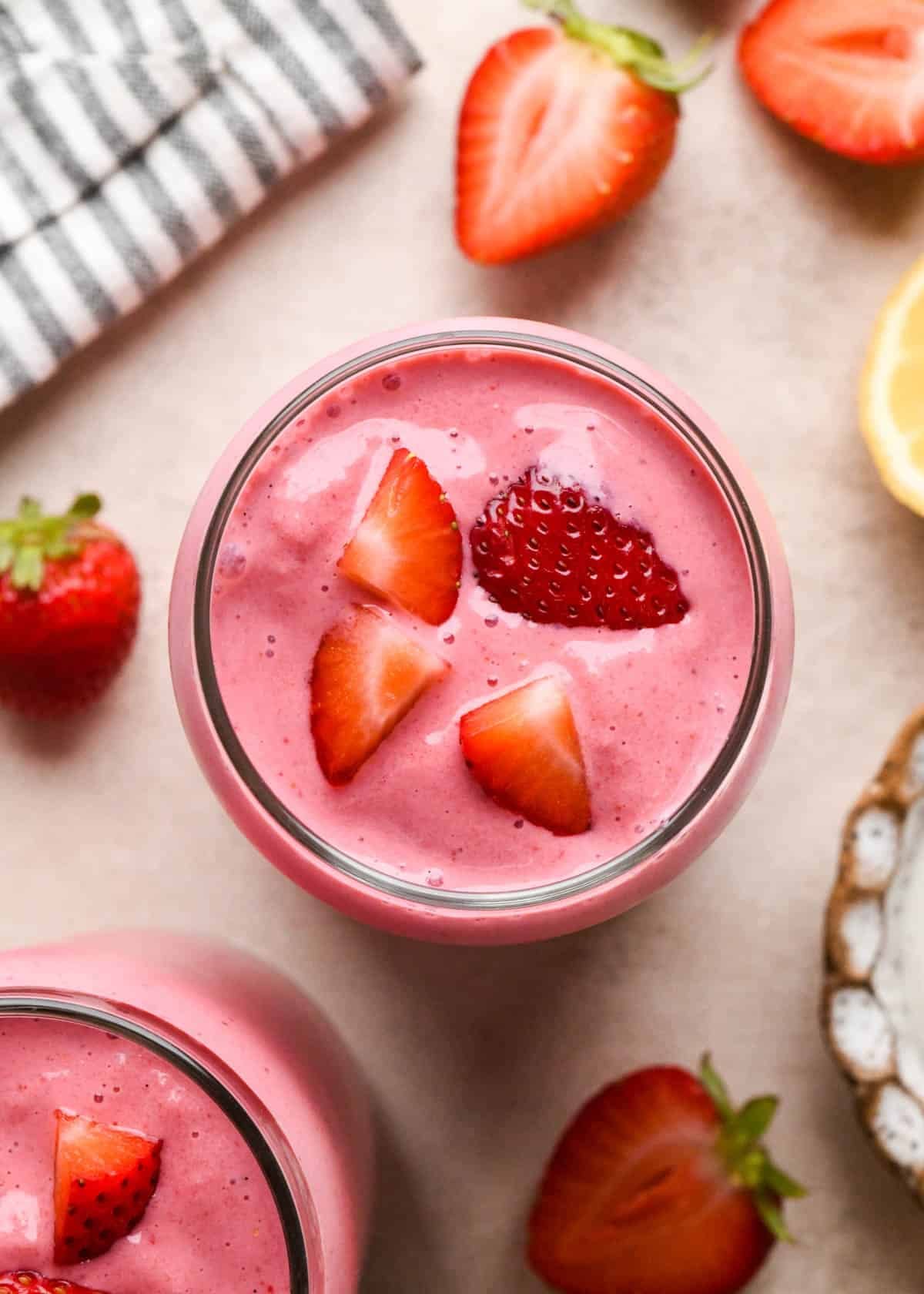 strawberry smoothie in a glass topped with chopped strawberries, surrounded by another glass of smoothie, strawberries and a lemon