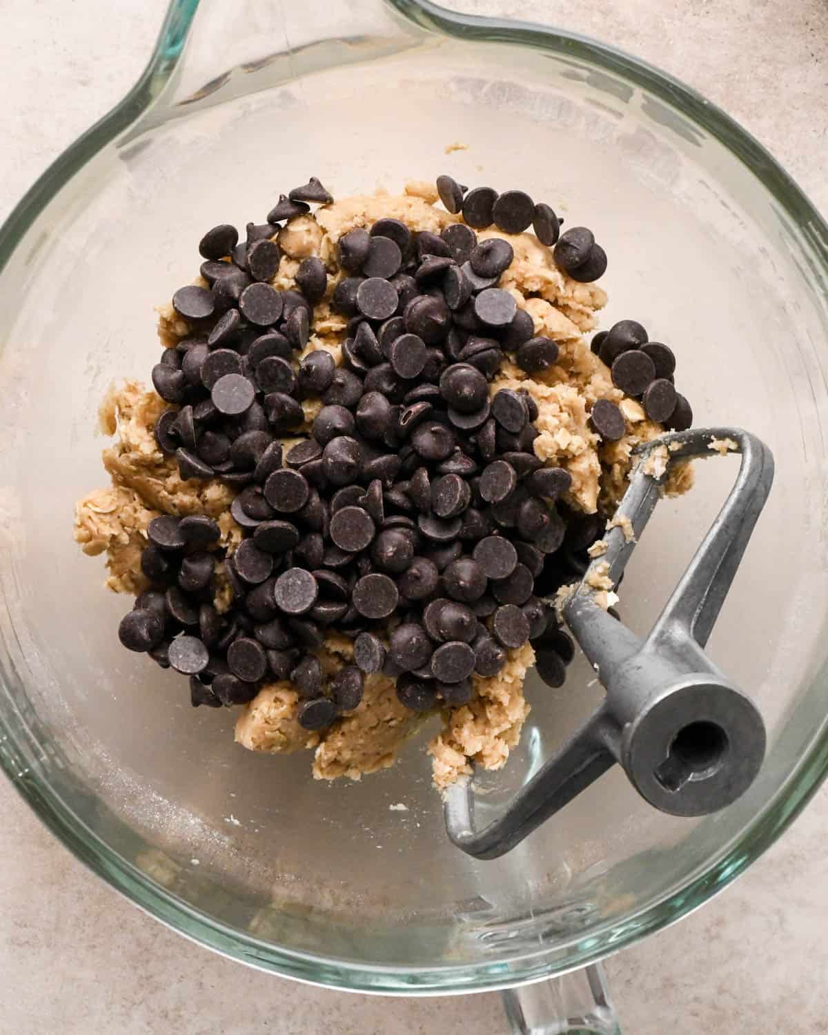 making vegan oatmeal cookies - adding chocolate chips to the dough in a glass mixing bowl with the paddle attachment, before mixing.