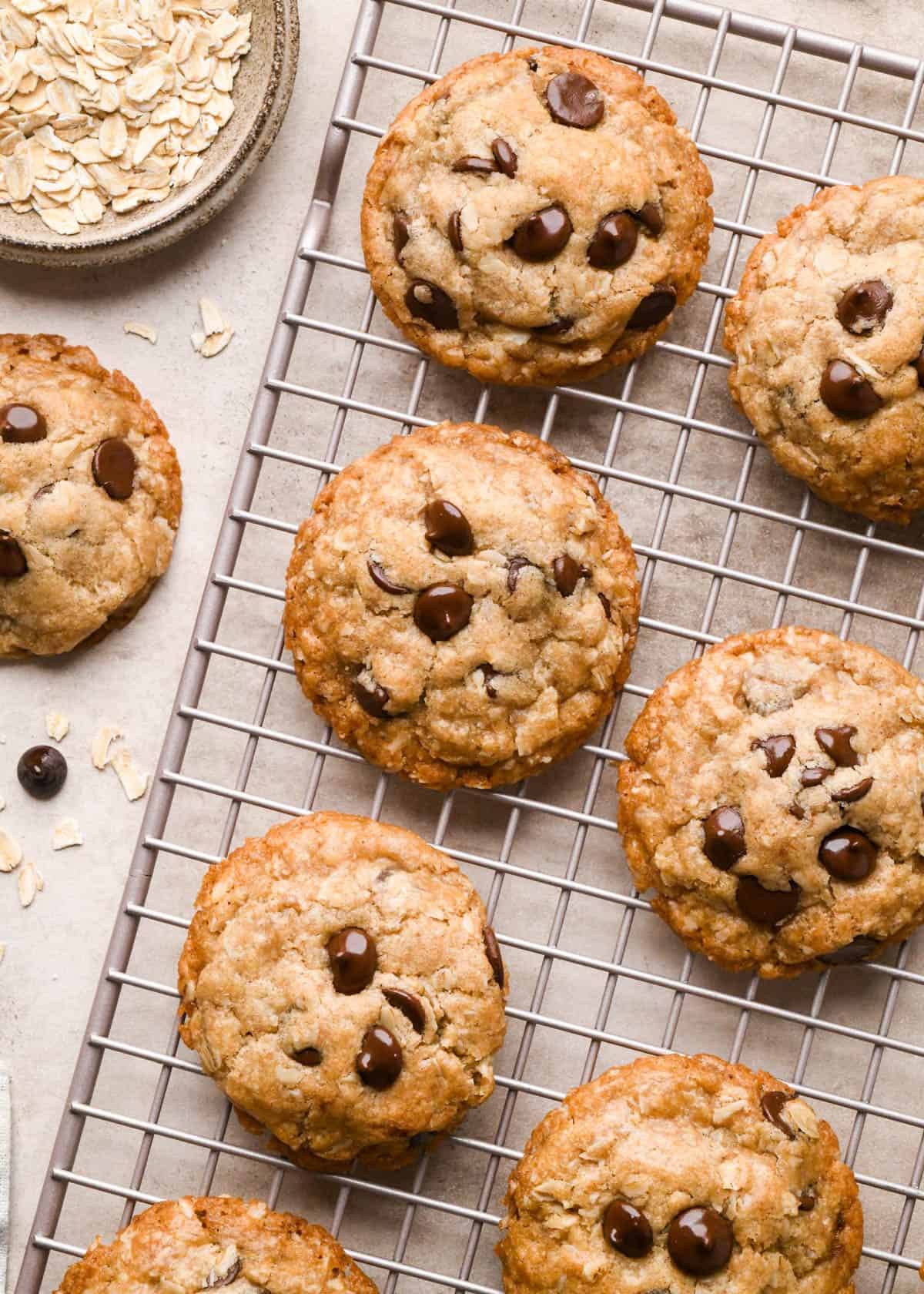 6 vegan oatmeal cookies with chocolate chips on a wire cooling rack with more cookies, oats and chocolate chips around it