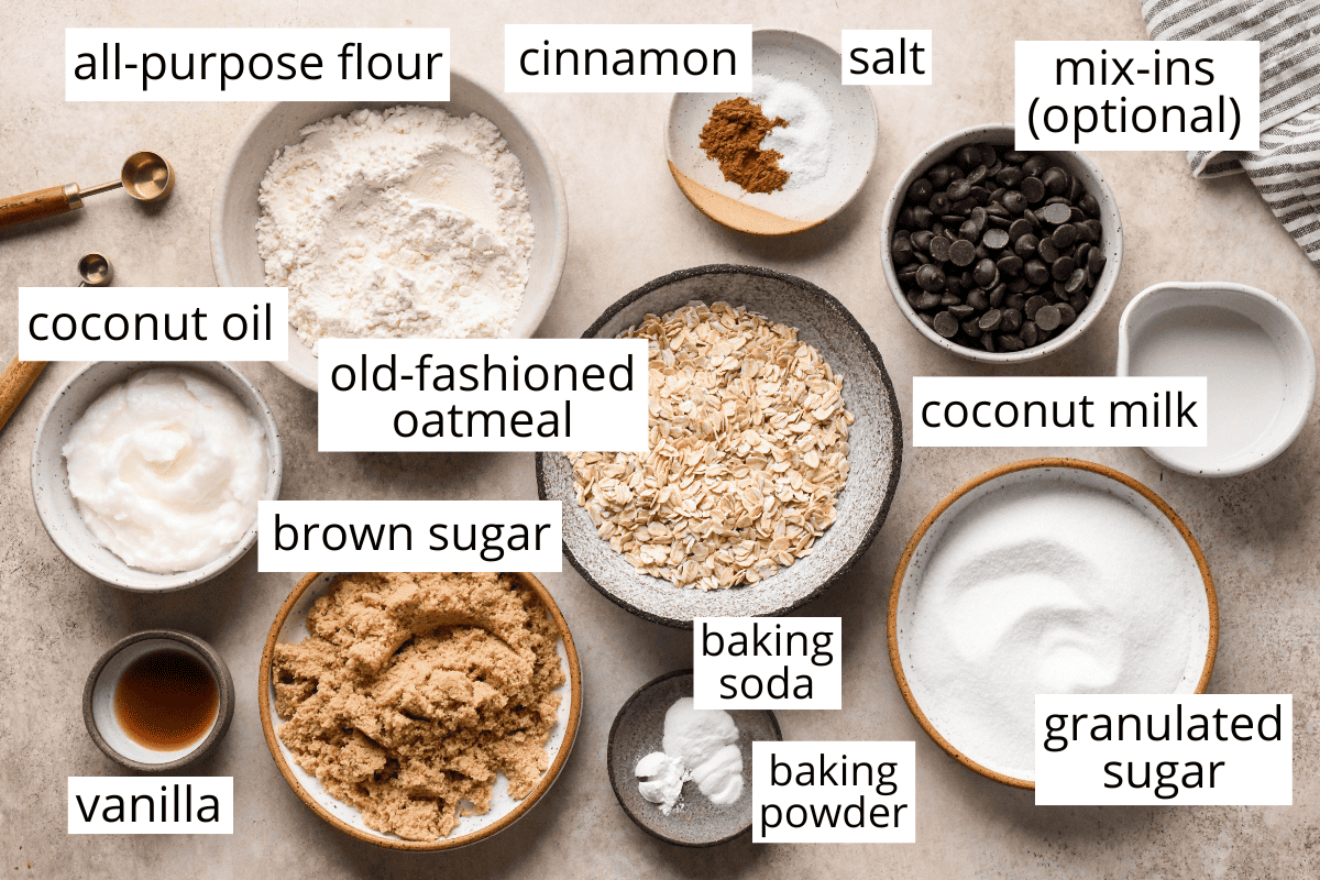 overhead photo of the ingredients in this vegan oatmeal cookie recipe, all in bowls and labeled like oatmeal, brown sugar, white sugar, coconut oil, coconut milk, chocolate chips and flour