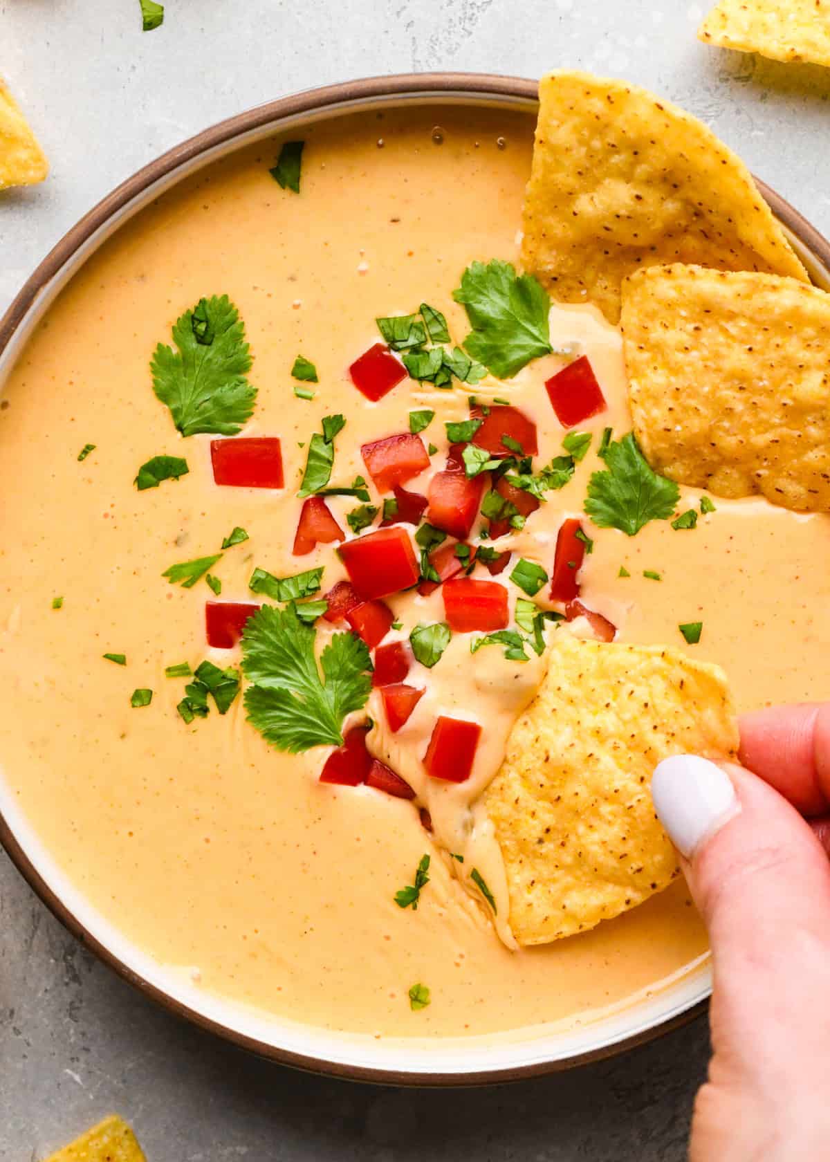 a hand dipping a tortilla chip into a Bowl of homemade queso dip topped with diced red peppers and chopped cilantro