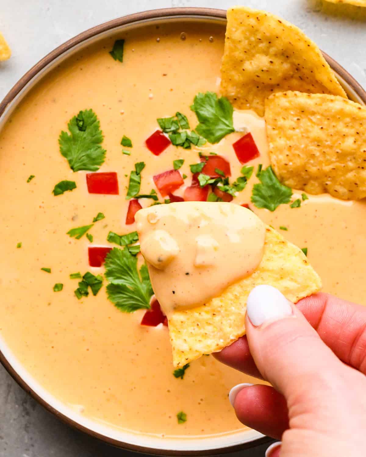 Bowl of homemade queso dip topped with diced red peppers and chopped cilantro, served with tortilla chips for dipping.