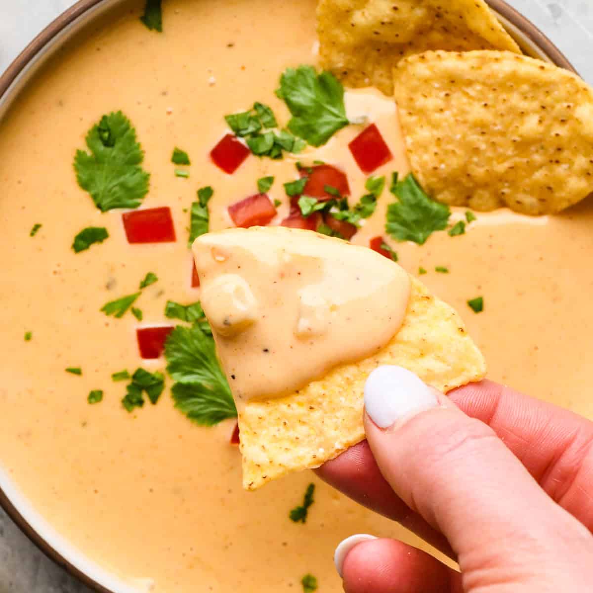 Bowl of homemade queso dip topped with diced red peppers and chopped cilantro, served with tortilla chips for dipping.