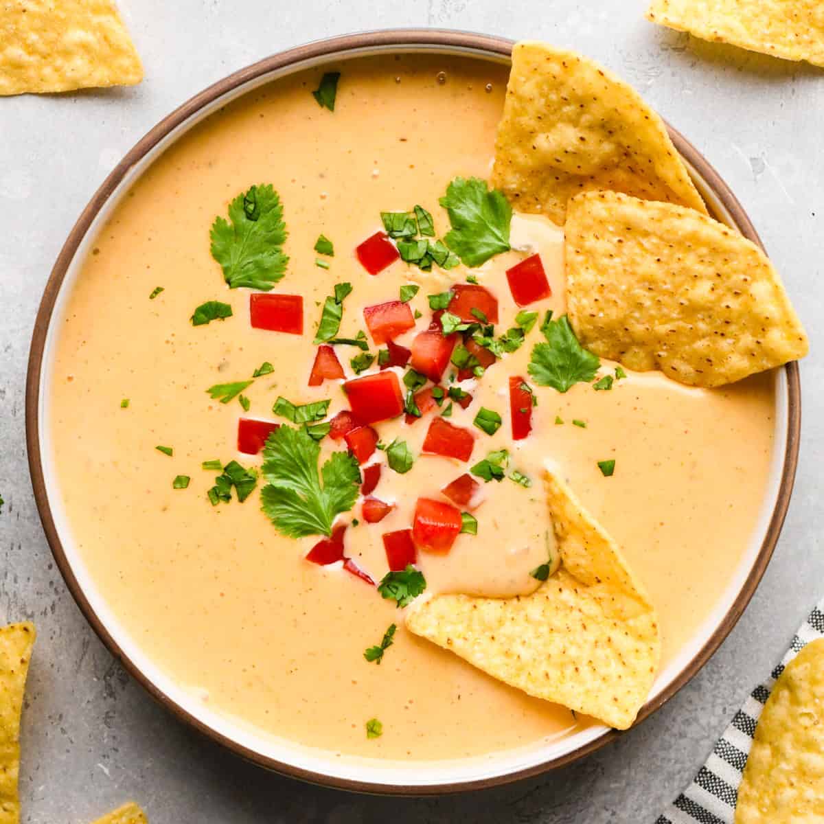 Bowl of homemade queso dip topped with diced red peppers and chopped cilantro, served with tortilla chips for dipping.