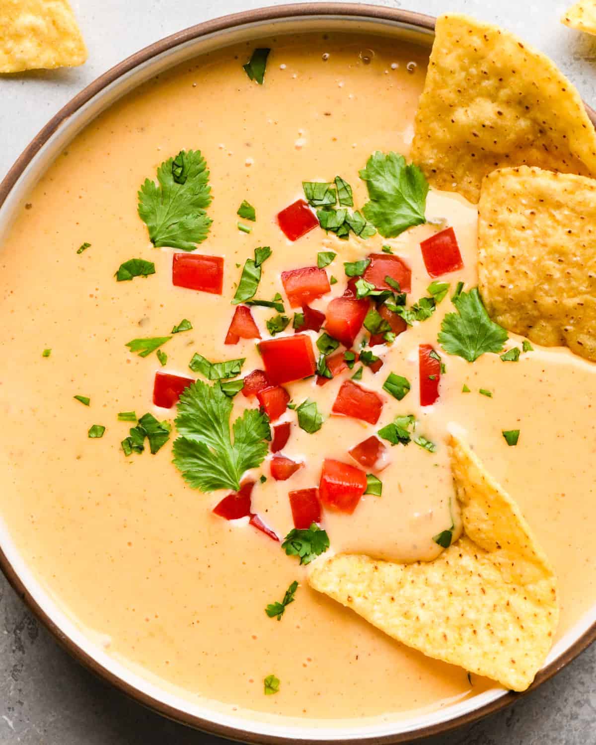 Bowl of homemade queso dip topped with diced red peppers and chopped cilantro, served with tortilla chips for dipping.