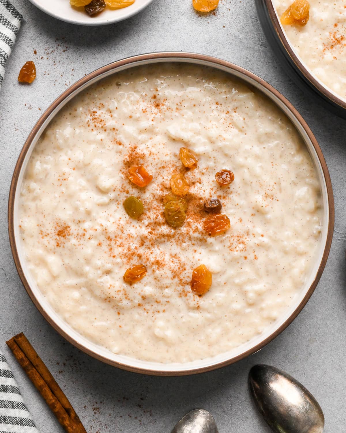 Overhead view of a bowl of creamy rice pudding topped with golden raisins and a dusting of cinnamon, with a spoon resting near the bowl.