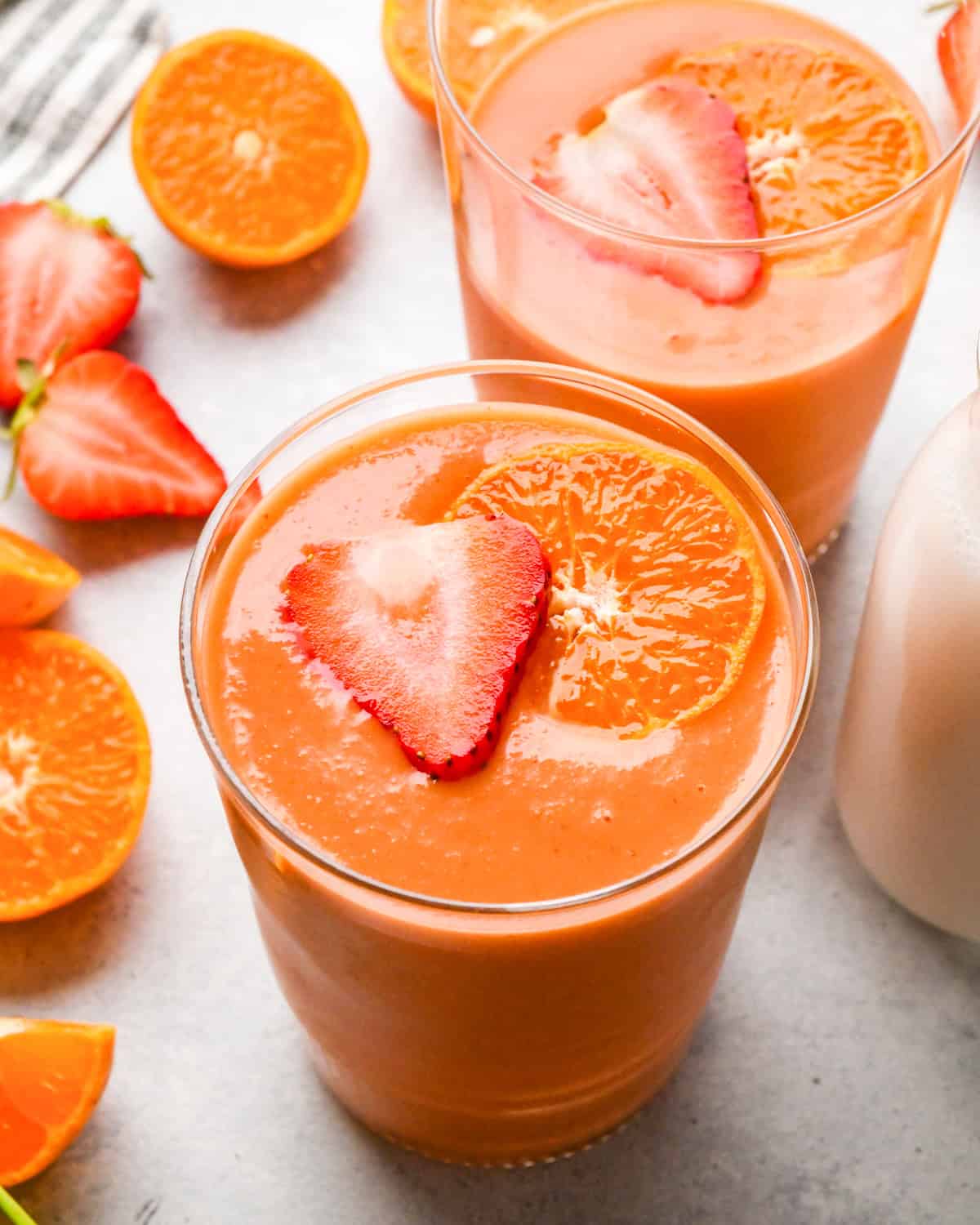 Overhead photo of 2 tall glasses of orange-colored carrot fruit smoothie garnished with strawberries and orange slices, with fresh fruit in the background.