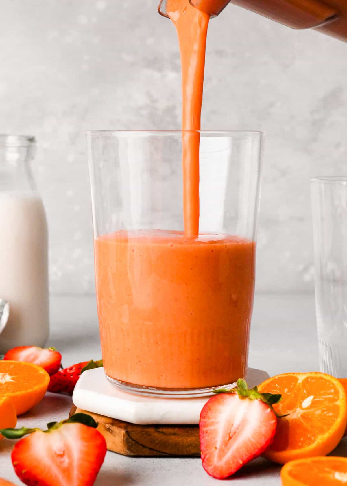 Making carrot fruit smoothie: front-view of completed smoothie being poured into a tall clear glass on a light surface. The glass is sitting on a white coaster and surrounded by slices of fruit and a jug of milk.