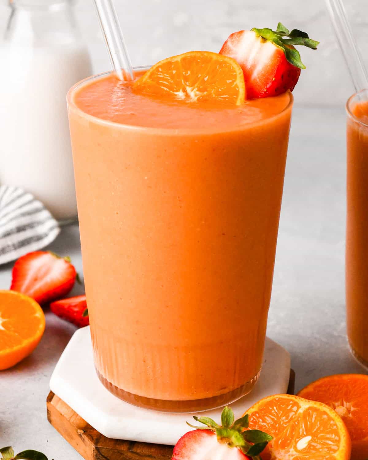 Tall glass of orange-colored carrot fruit smoothie garnished with a strawberry and an orange slice, with a straw in the glass and fresh fruit in the background.
